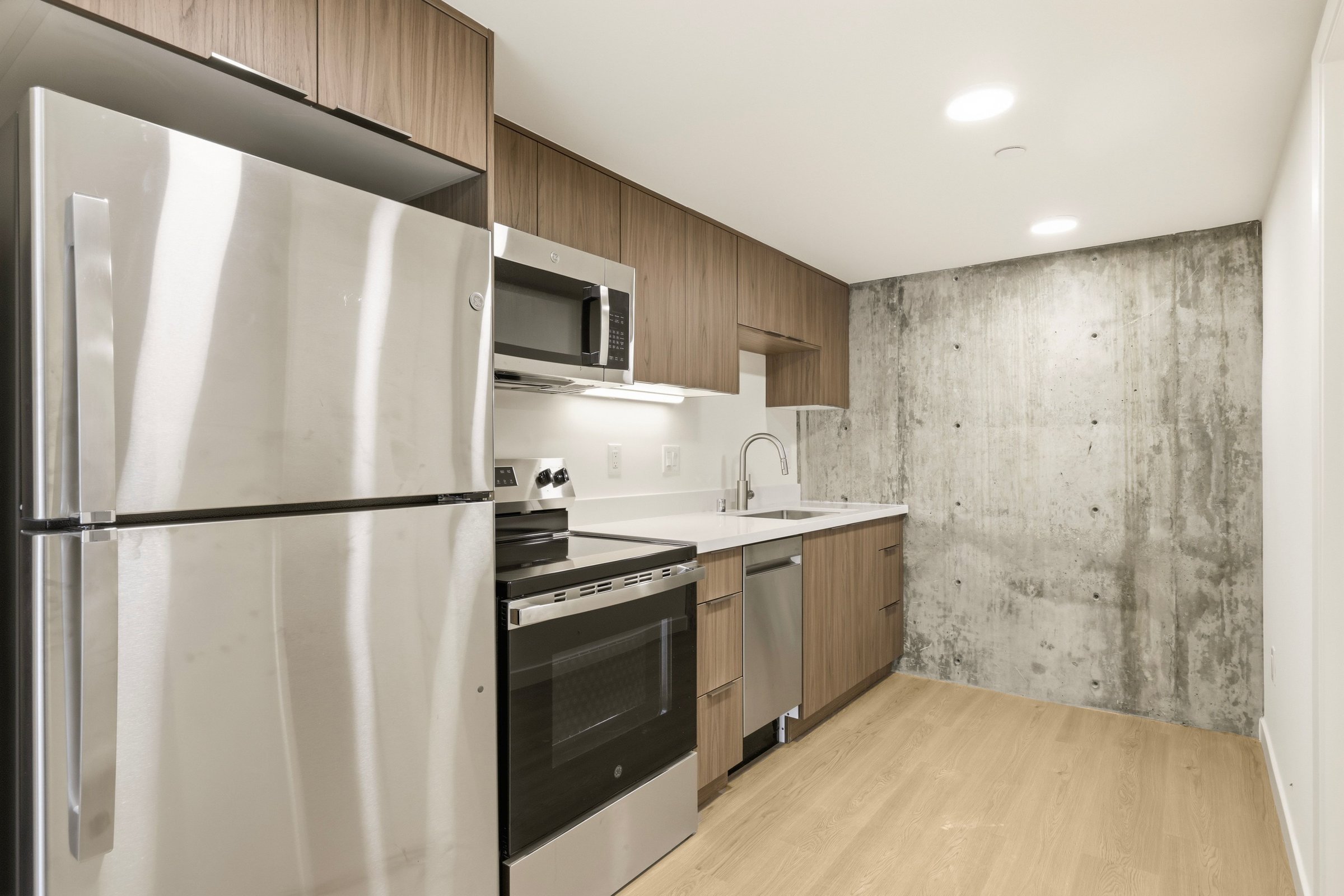 Goldilocks apartment kitchen with stainless steel appliances, wood cabinetry, and a concrete accent wall.