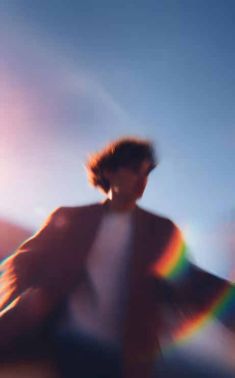 Blurred person outdoors with curly hair against blue sky and rainbow light flare.
