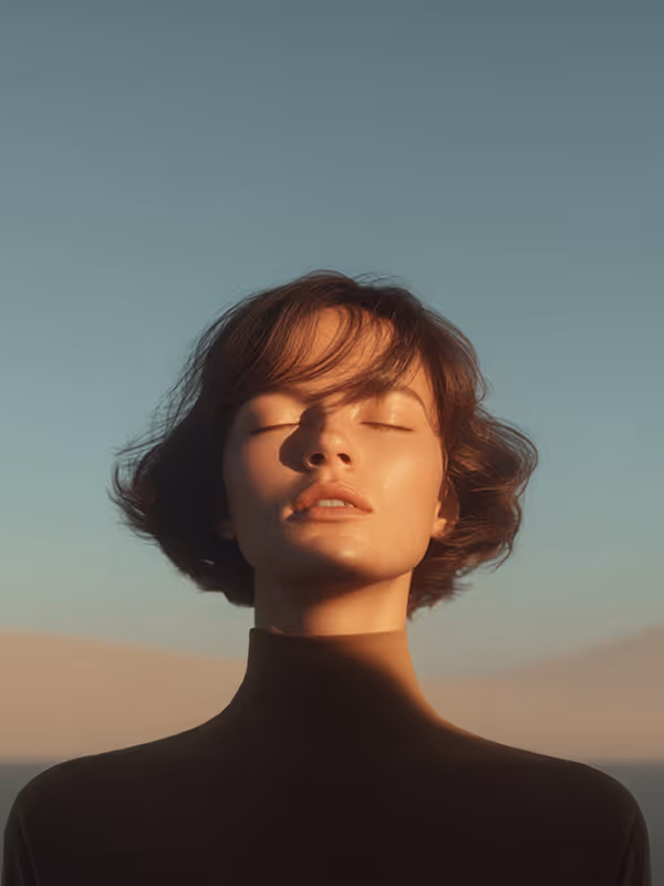 Woman with short hair and closed eyes facing sunlight against a clear blue sky.
