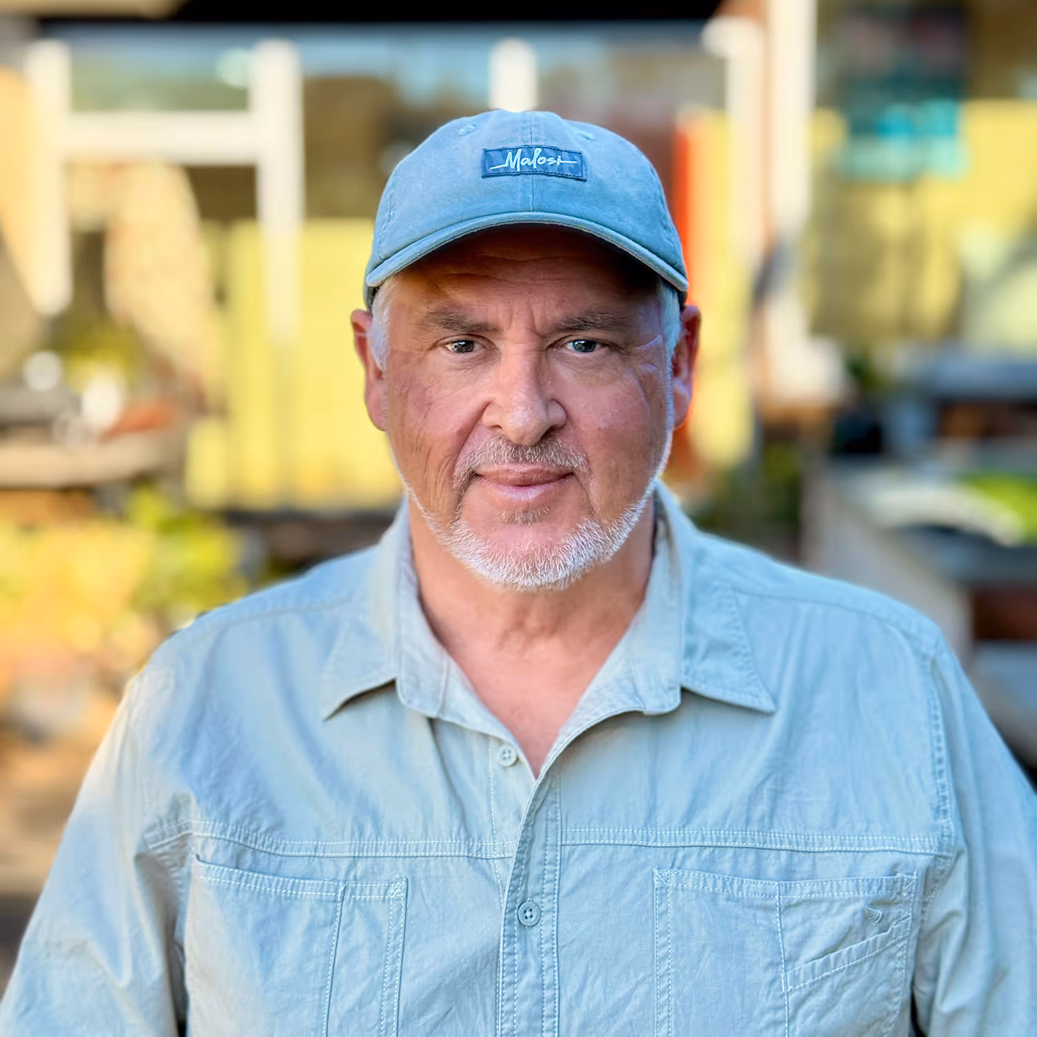 Middle-aged man with a white beard wearing a blue cap and light blue shirt, outdoors in a blurred background.