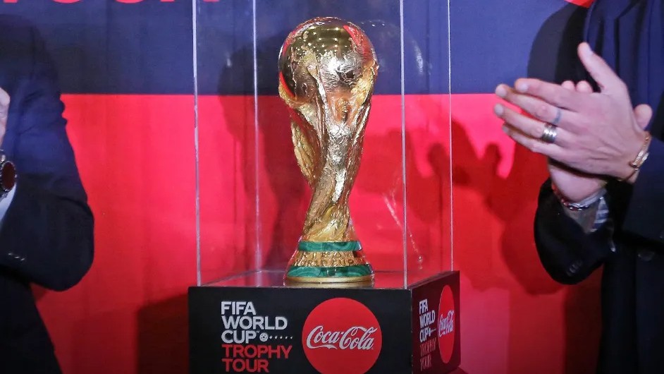 A golden FIFA World Cup trophy, with a green base, is displayed in a clear case on a black pedestal featuring "FIFA WORLD CUP TROPHY TOUR" and "Coca-Cola" logos. A vibrant red and dark blue backdrop is behind it. On the left, a person's arm with a watch is visible, while on the right, hands in a dark suit, adorned with rings and a bracelet, are seen.