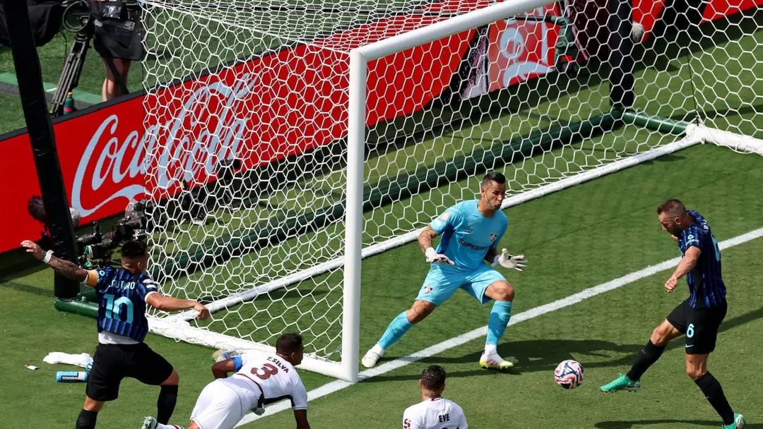 A soccer player in a dark blue striped jersey kicks a multi-colored ball towards a white goal with a hexagonal net. A goalkeeper in a light blue uniform is positioned defensively, while other players in white and striped jerseys are near the goal. A red Coca-Cola sign is visible behind the net on the bright green field under clear daylight.