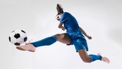 A female soccer player in a blue uniform is captured mid-air, powerfully kicking a black and white soccer ball with her left foot. She wears a blue jersey, shorts, and socks, along with light-colored athletic shoes. The background is a plain, bright white, and the scene is evenly lit.