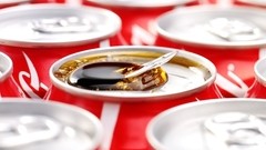 A close-up view shows numerous red soda cans with silver lids, arranged in a pattern. One central can is open, revealing dark brown cola and a small silver spoon resting within the liquid. Bright, even lighting illuminates the scene, creating reflections on the metallic surfaces.