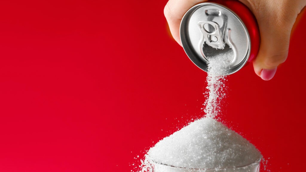 A human hand with pink nail polish holds an open red soda can, pouring a cascade of white granulated sugar into a clear glass, forming a large mound. The entire scene is brightly lit against a solid, vivid red background.
