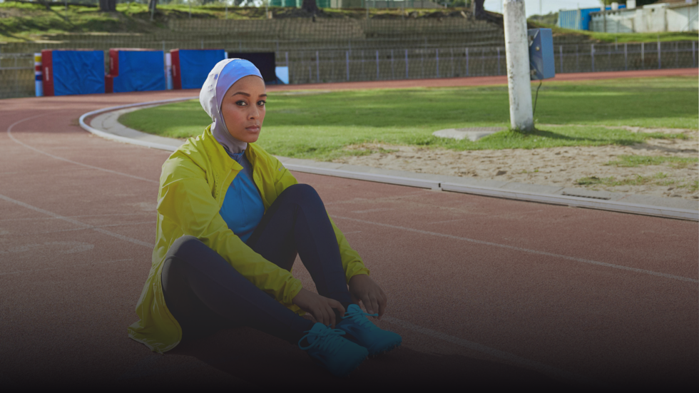 A woman in a yellow jacket and blue hijab sits on a reddish-brown running track, tying her teal shoes. She looks towards the viewer, with a green field and sand pit in the background under bright daylight.