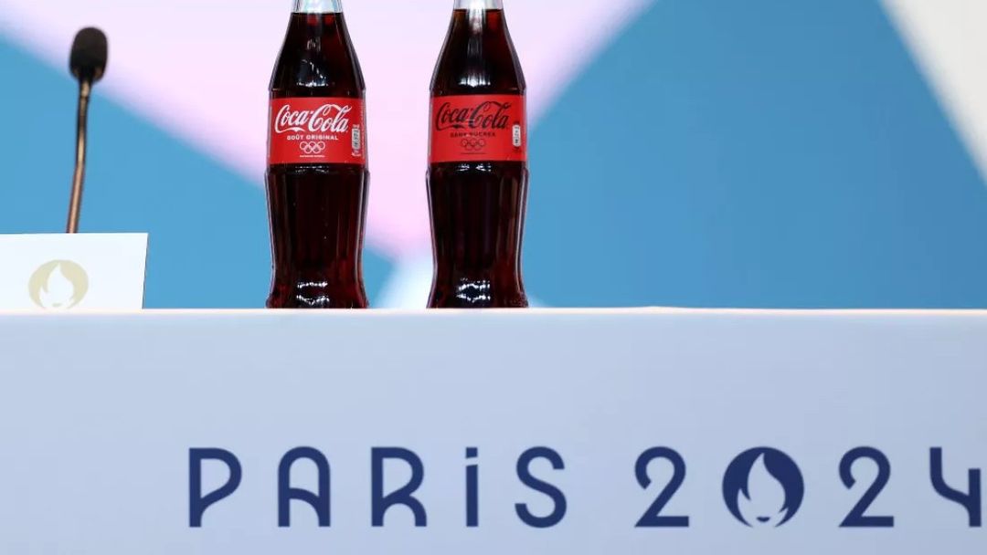Two dark Coca-Cola bottles with red labels, one marked 'GOÛT ORIGINAL' and the other 'SANS SUCRES', stand on a white surface. A black microphone and a white card with a gold emblem are to the left. The white surface features the dark blue text 'PARIS 2024', with the '0' represented by the Paris 2024 emblem. The background is a light blue wall, illuminated by bright, even lighting.