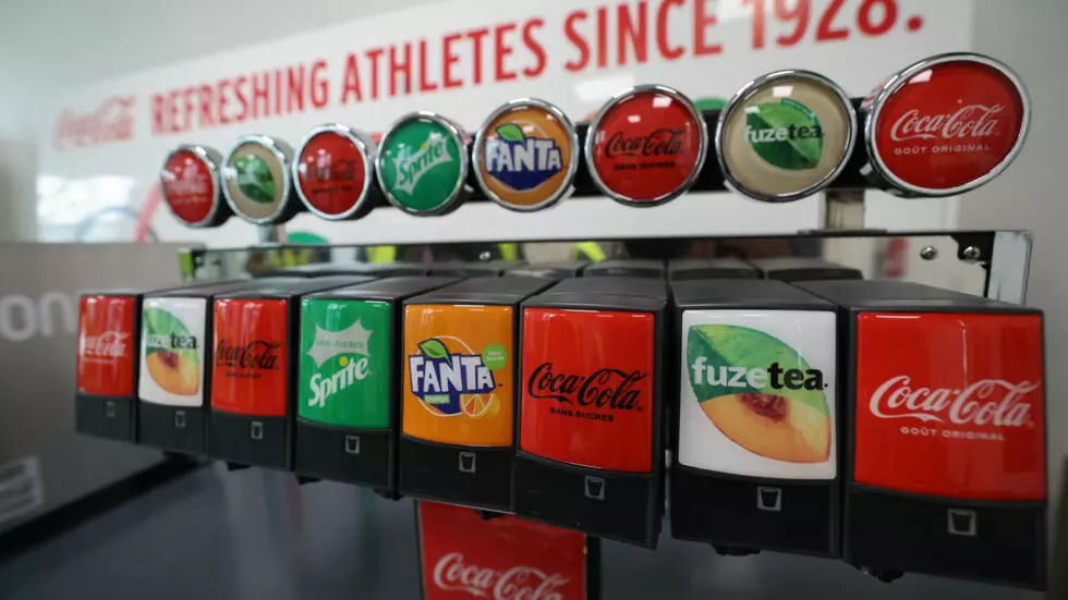 A brightly lit beverage dispenser features a row of eight spouts, each labeled with a distinct drink brand. Above the spouts, corresponding circular brand emblems are arranged on a metallic bar. Visible brands include Coca-Cola (original and sugar-free), Sprite sugar-free, Fanta, and Fuze Tea peach. The dispenser units are black, showcasing vibrant red, green, orange, and white labels. A white sign with red lettering, partially visible in the background, states 'Coca-Cola REFRESHING ATHLETES SINCE 1928.'