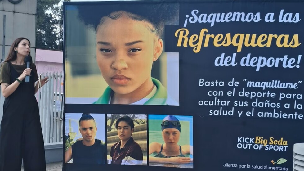 A woman with long dark hair, wearing a dark green shirt and black jumpsuit, speaks into a black microphone while standing next to a large banner. The banner features a serious young Black woman's face prominently, with three smaller portraits of other young people below. Large Spanish text on the dark banner reads "¡Saquemos a las Refresqueras del deporte!" and "Basta de 'maquillarse' con el deporte para ocultar sus daños a la salud y el ambiente," alongside the English logo "KICK Big Soda OUT OF SPORT." A light pink building and white fence are visible in the background under diffused daylight.