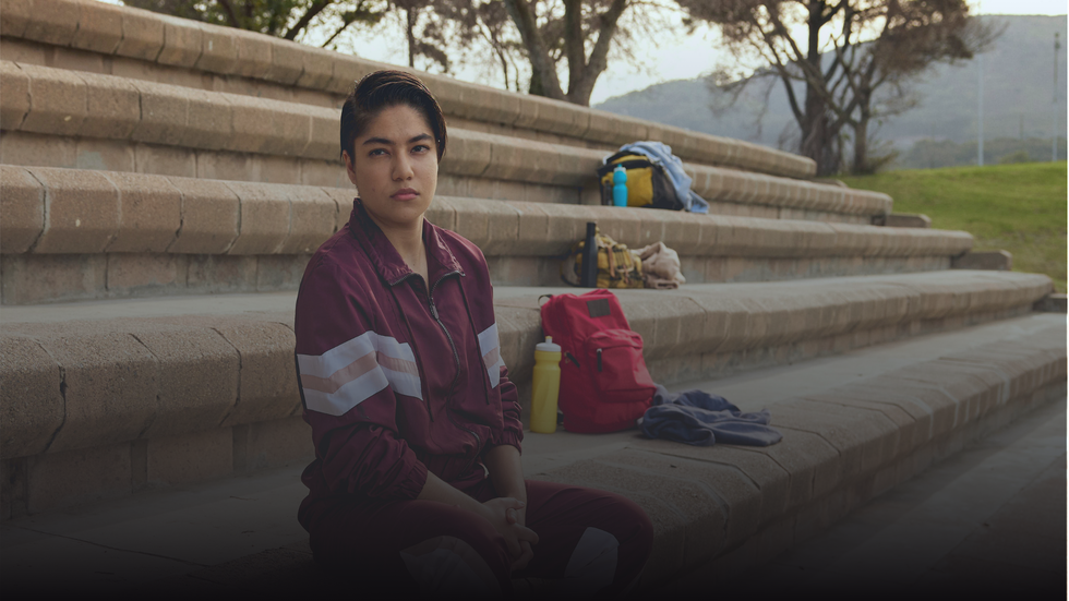 A person with short dark hair and a serious expression sits on a light brown stone staircase, wearing a maroon tracksuit with white and peach stripes. Various bags, water bottles, and clothing items are placed on the steps behind them. The background features a grassy hill, bare trees, and distant mountains under a soft, natural light.