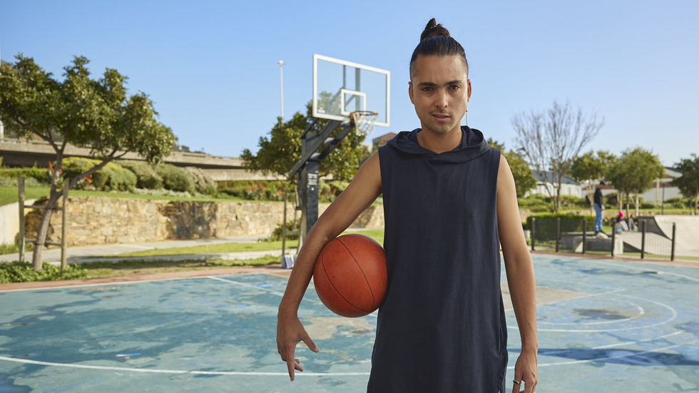 A young man with a man bun and a serious expression stands on a blue basketball court, holding an orange basketball under his left arm. He wears a dark blue sleeveless hooded top. In the background, a basketball hoop is visible on the left, and a skate park with blurred figures is on the right. The court is surrounded by green trees, a stone wall, and distant buildings under a clear blue sky, illuminated by bright daylight.
