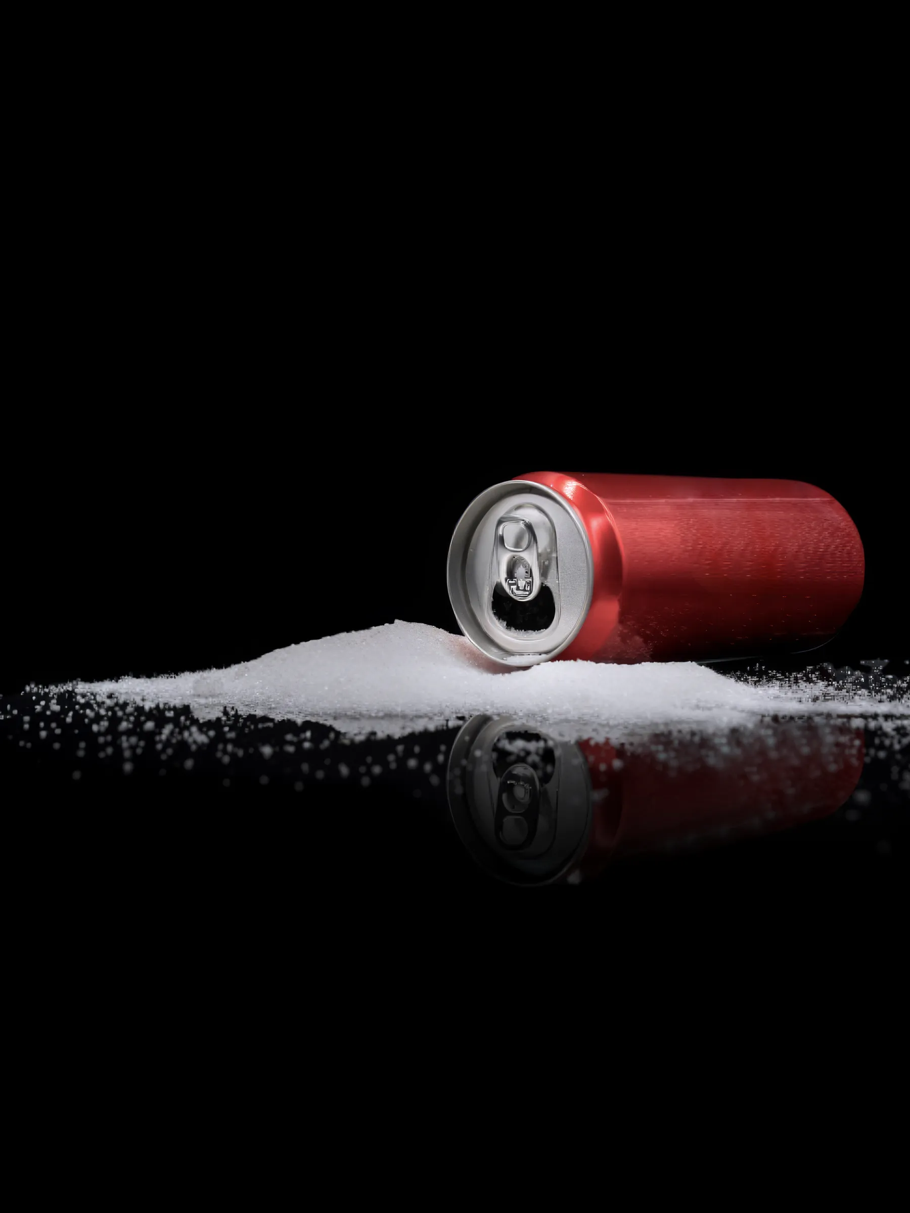 A red aluminum soda can rests horizontally on a pile of white granulated sugar, with its opening facing left. The objects are brightly illuminated against a deep black background, creating a clear reflection on the glossy surface below.