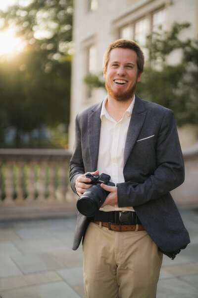 Smiling man in a blazer holding a camera outdoors in front of a building at sunset.