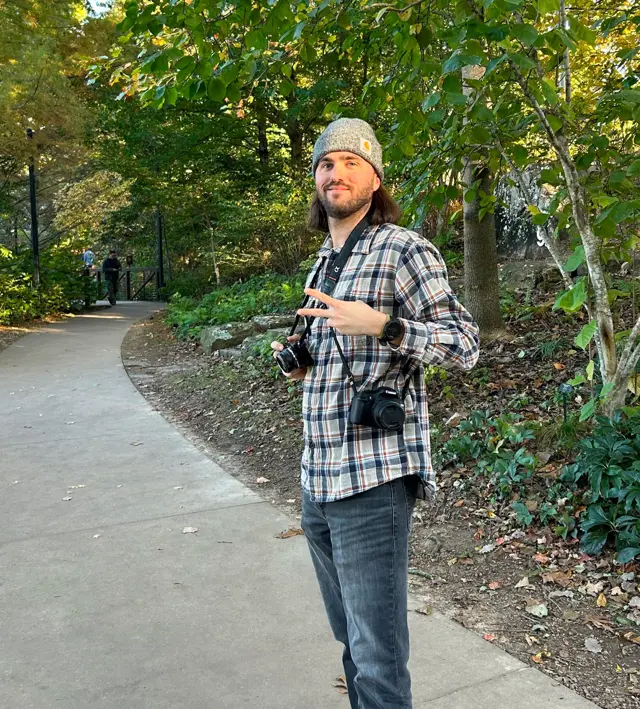 Man in a gray beanie and plaid shirt holding cameras while standing on a park pathway with greenery.