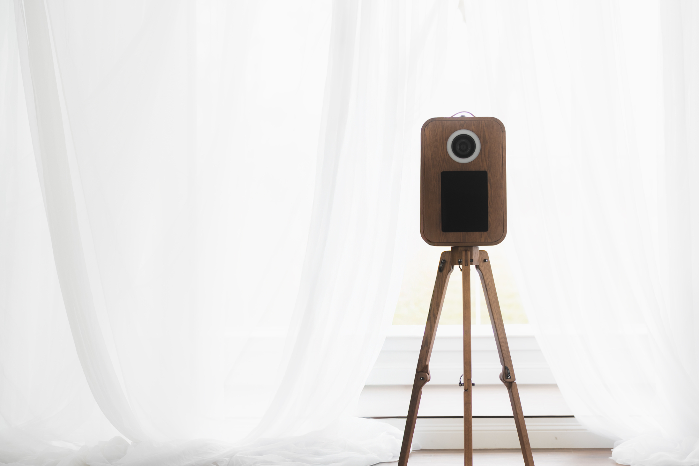 Wooden photo booth on a tripod stand in front of sheer white curtains and a window.