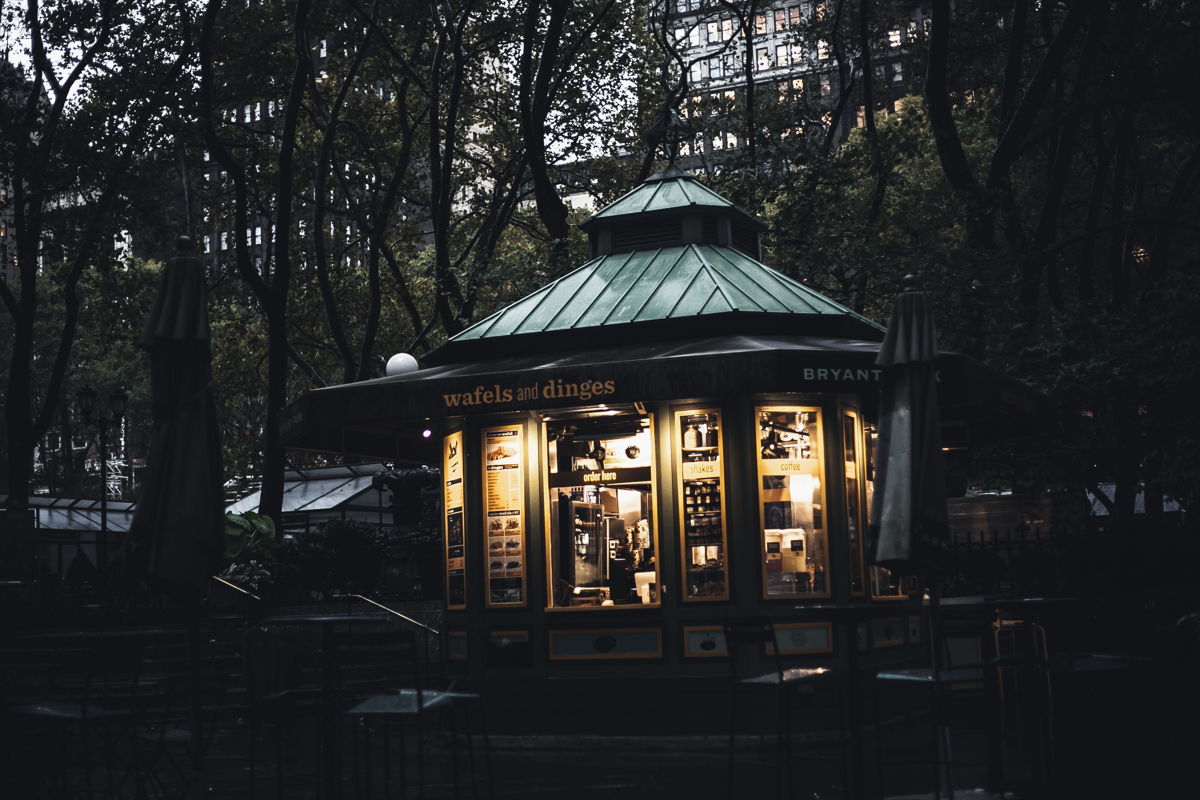 A warmly lit food kiosk glows at dusk among the trees of Bryant Park, with Midtown towers rising behind. Photo by Keenan Constance on Pexels.