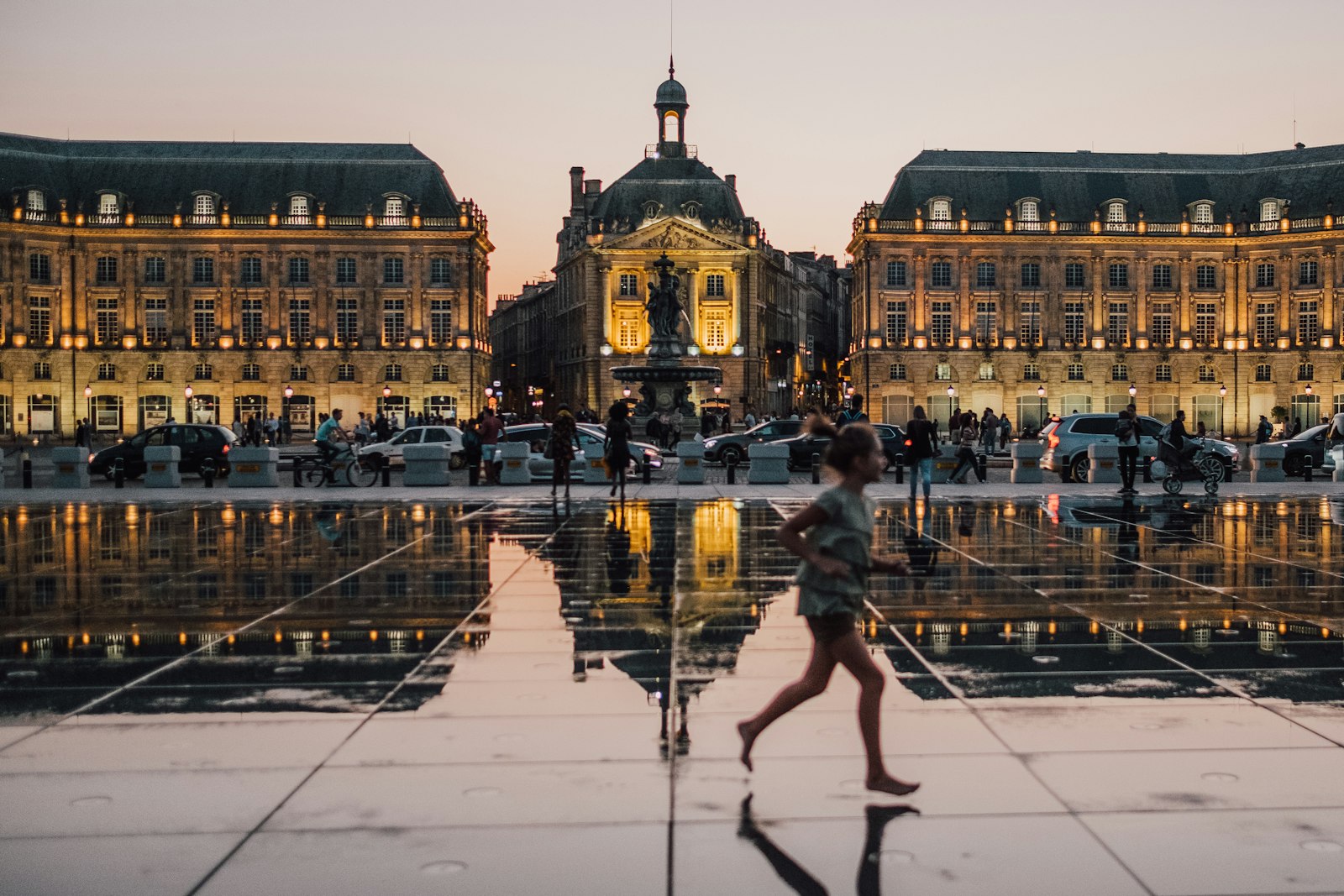 La Place de la Bourse reflejada en el estanque del Miroir d'eau al atardecer en Burdeos, con una figura cruzando el agua. Foto de Guillaume Flandre en Unsplash.