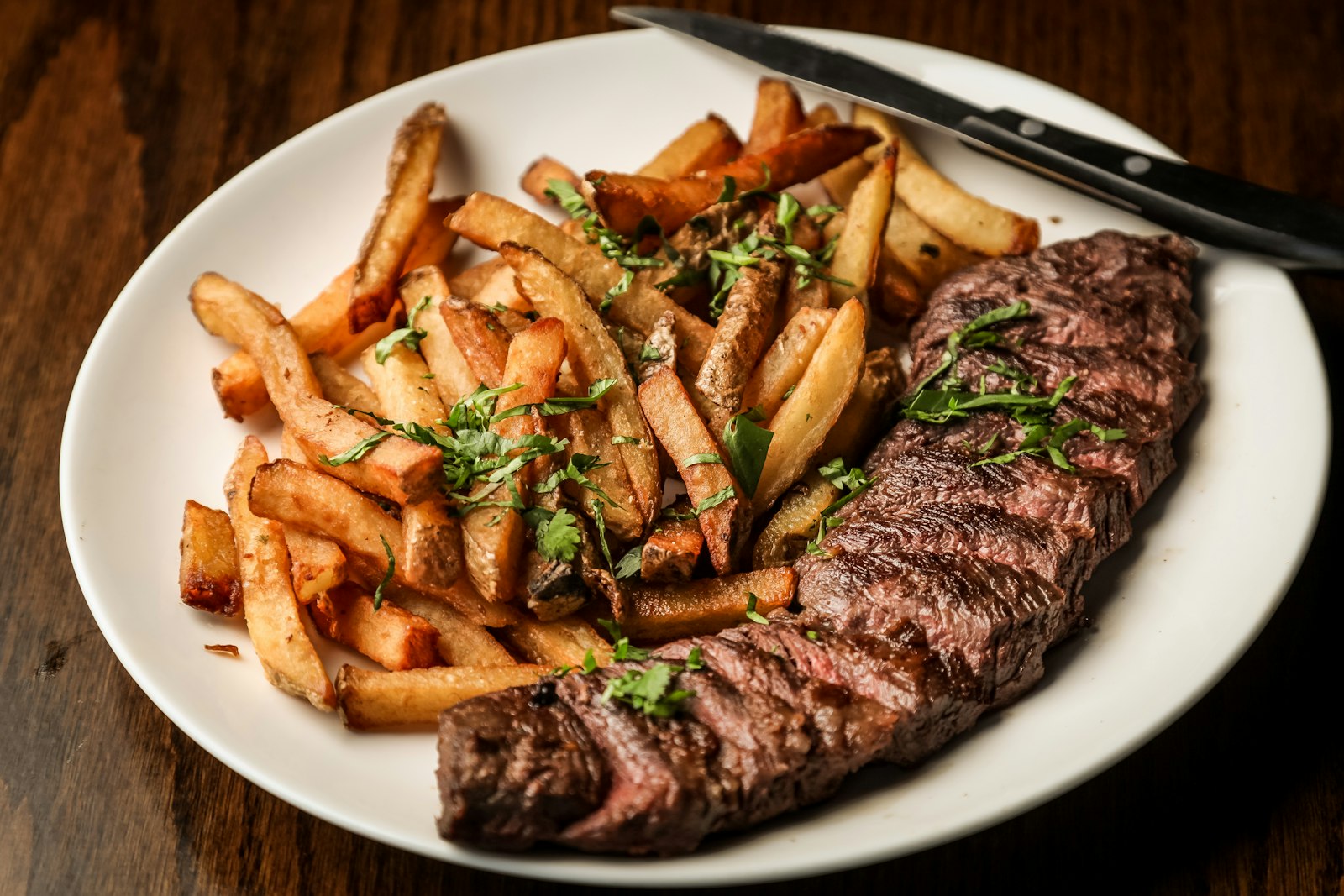 Un bife argentino a la parrilla con una montaña de papas fritas y hierbas en un plato blanco. Foto de Tim Toomey en Unsplash.