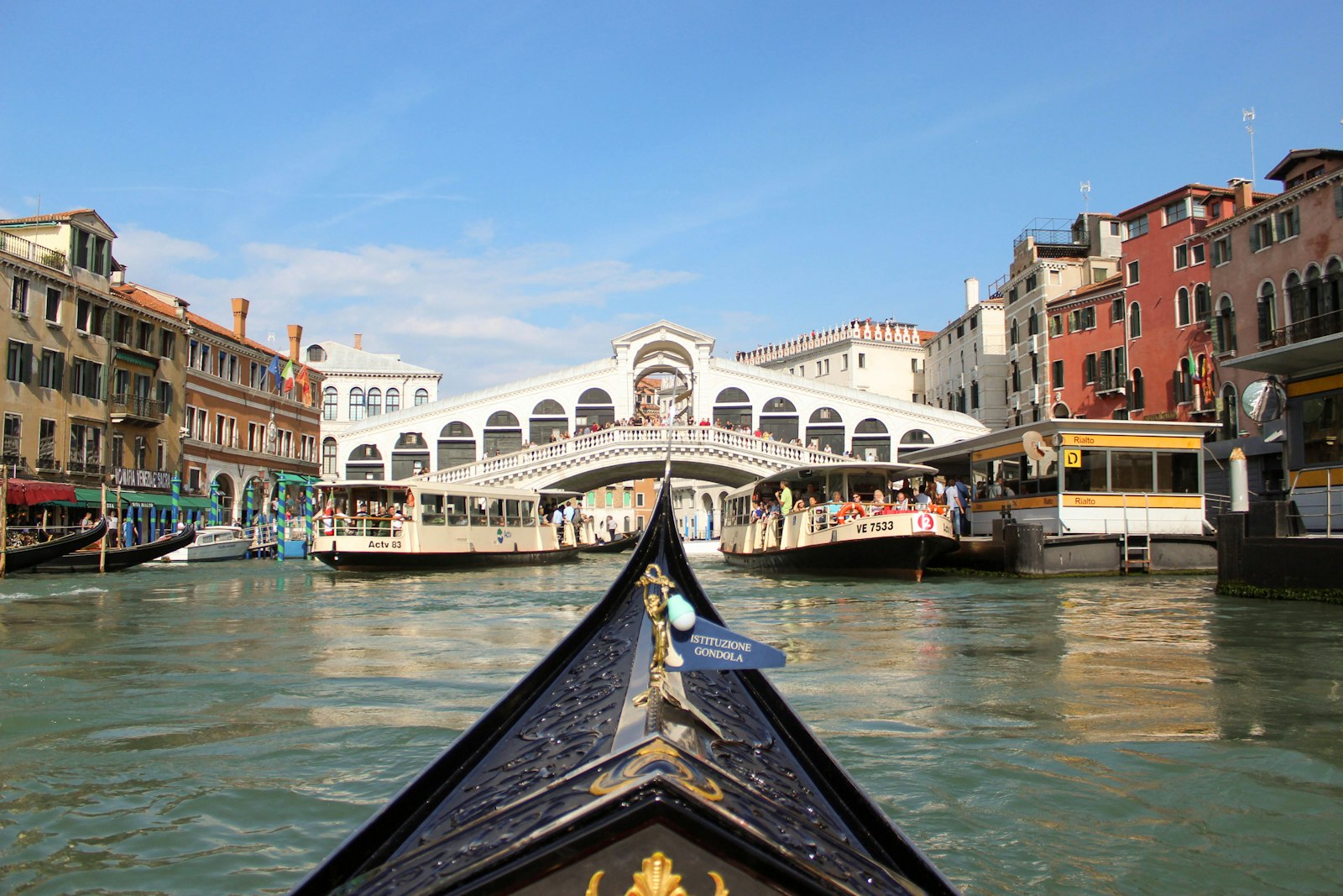 A gondola prow framing two crowded vaporetti and the Rialto Bridge on Venice's Grand Canal. Photo by KaLisa Veer on Unsplash.