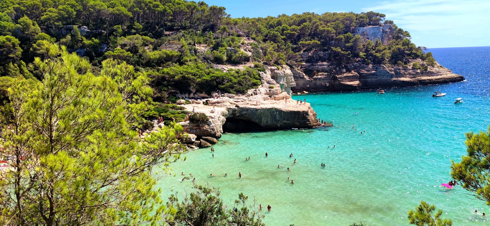 A group of friends swimming in the turquoise water of a pine-backed cove on Menorca. Photo by Fernando Jimenez on Unsplash.