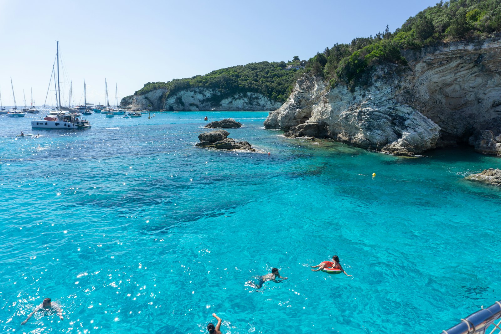 Sailboats anchored in a clear turquoise cove while swimmers float below the cliffs. Photo by Renaldo Kodra on Unsplash.