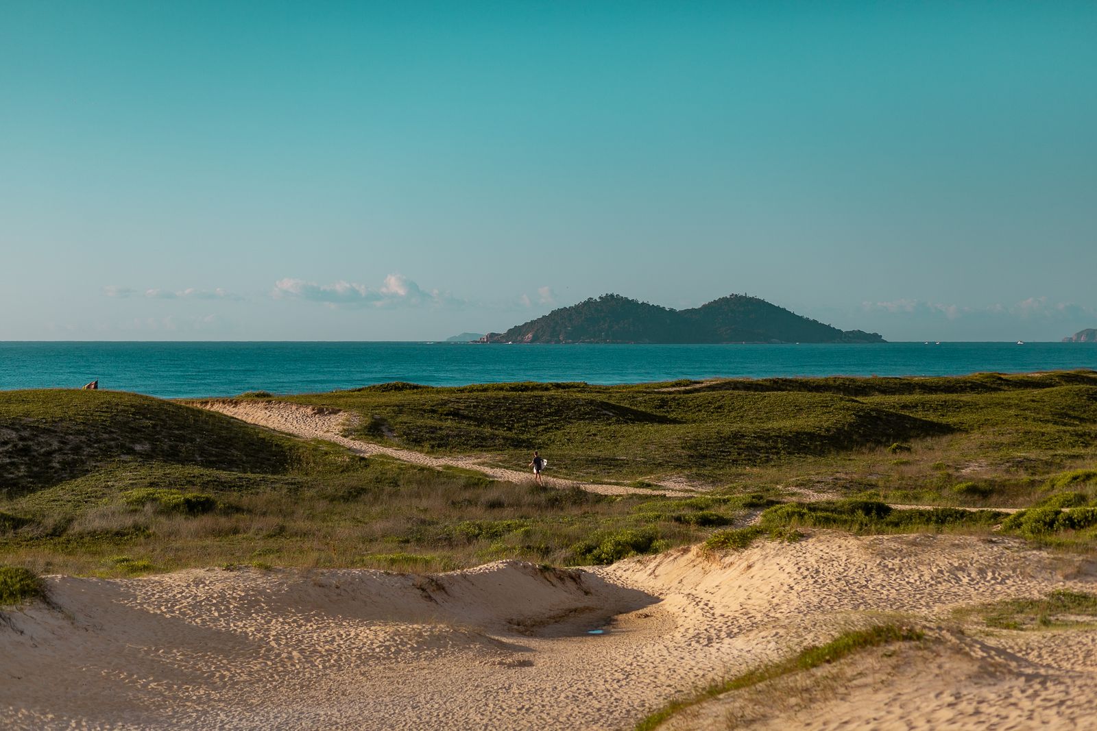 Sand dunes and open ocean with a distant island offshore, on the coast of Florianópolis. Photo by Clayton de Araujo on Pexels.