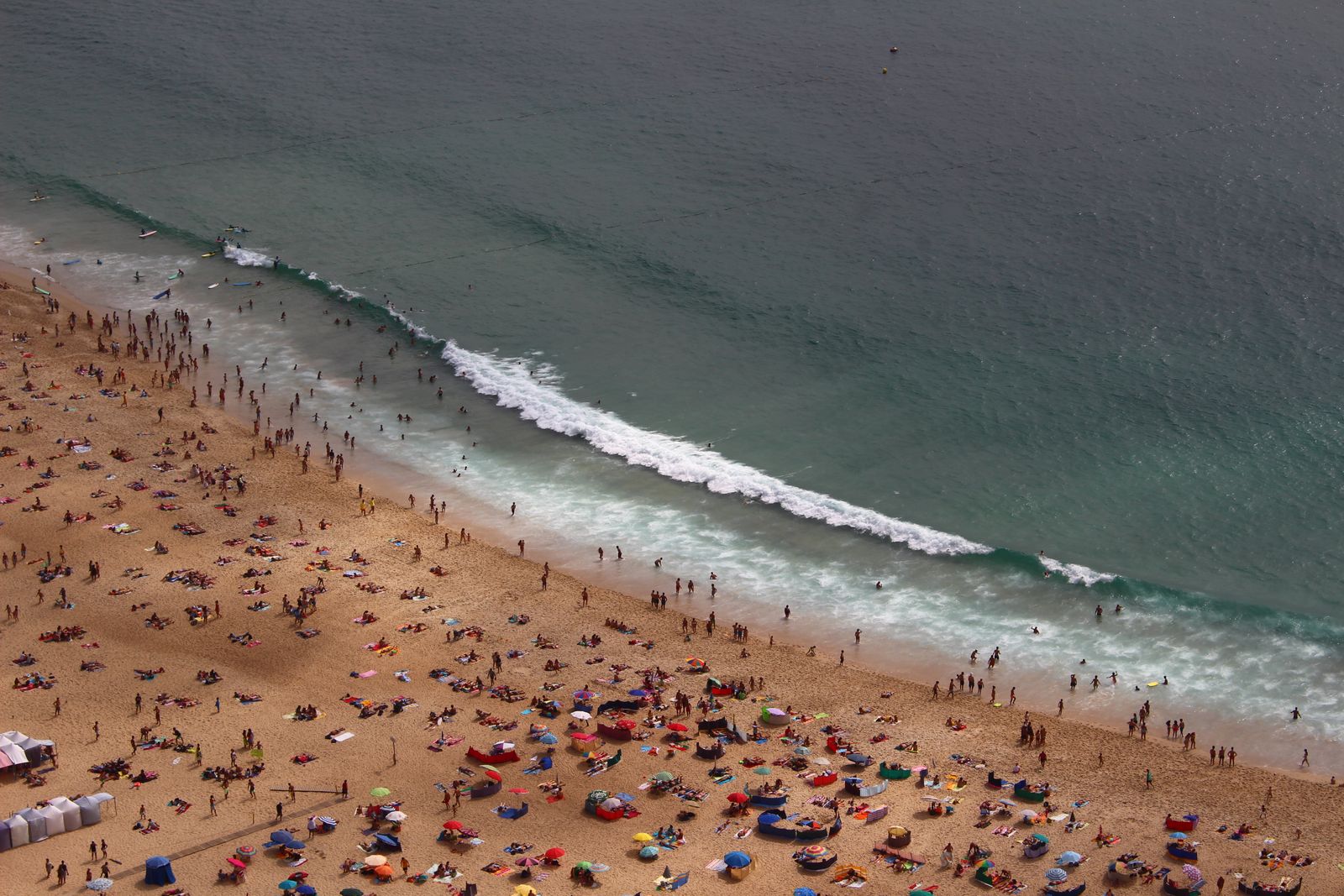 An aerial view of a packed summer beach, umbrellas and swimmers filling the sand and shallows. Photo by Mariana Vale on Pexels.