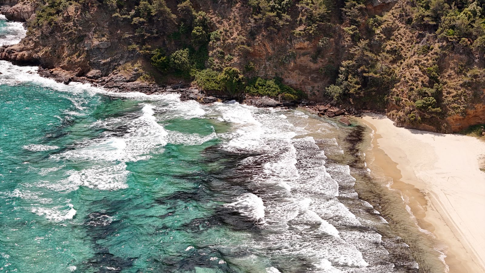 Turquoise water breaking over rocks beside a pale curve of sand, seen from above. Photo by Daniel Lepădatu on Pexels.