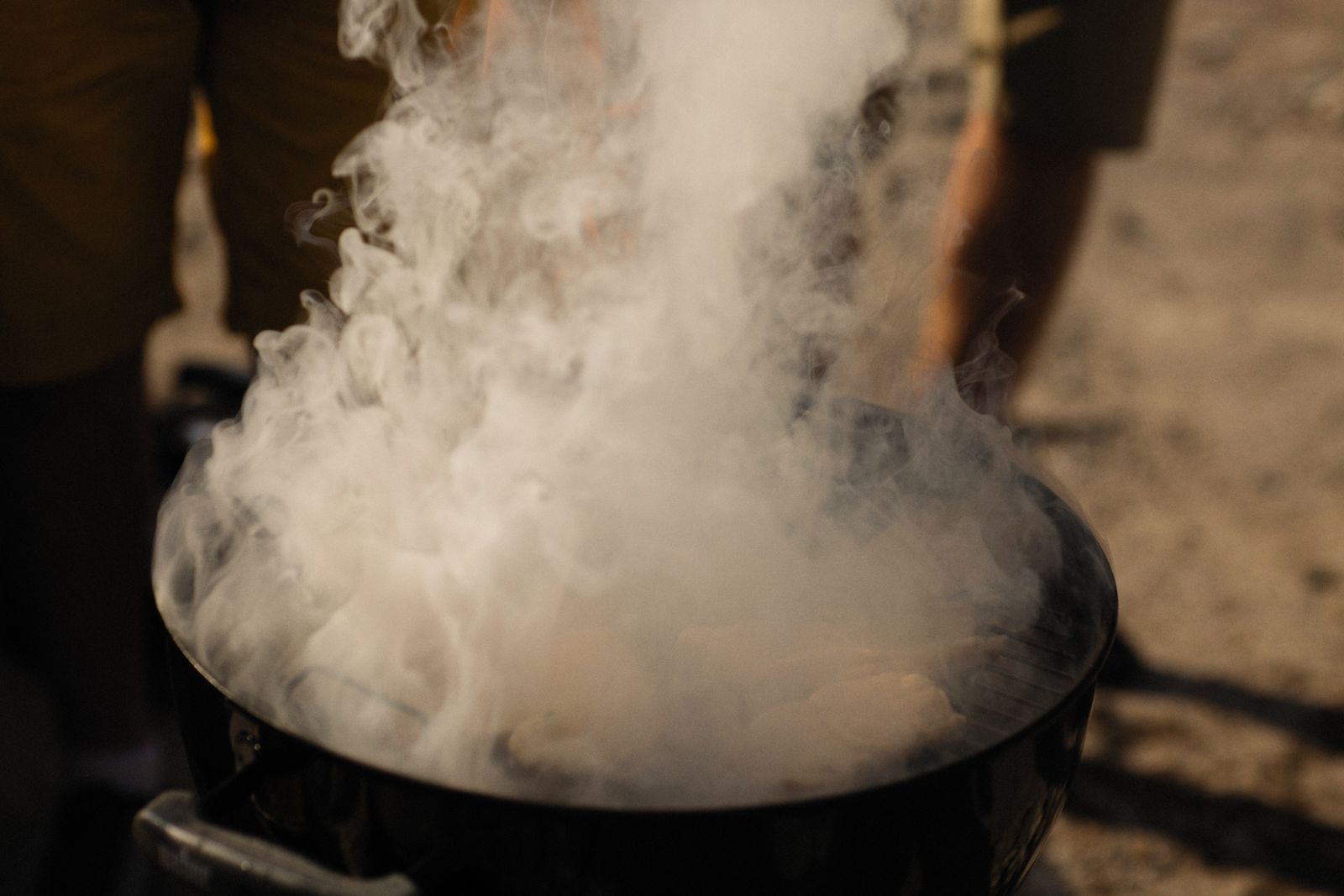 A charcoal grill smoking at an outdoor barbecue with people gathered round it. Photo by Alexey Demidov on Pexels.