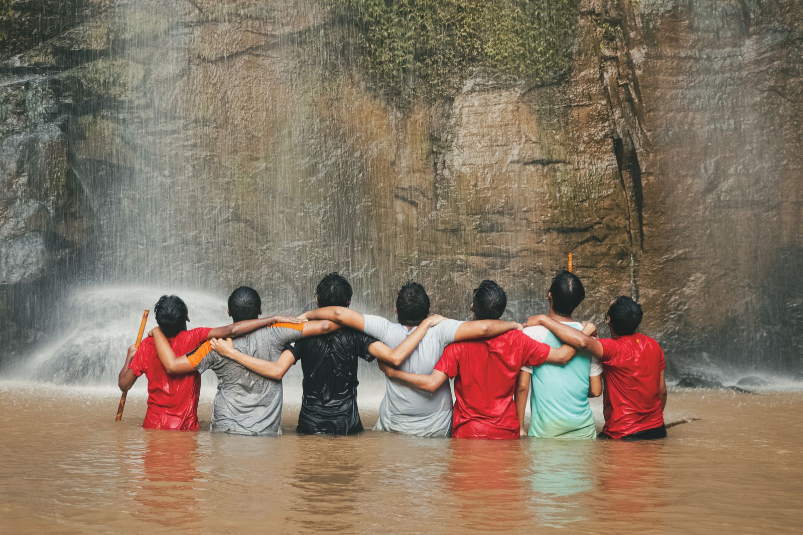 Friends standing arm in arm in the river below a waterfall. Photo by Ferdous Hasan on Pexels.