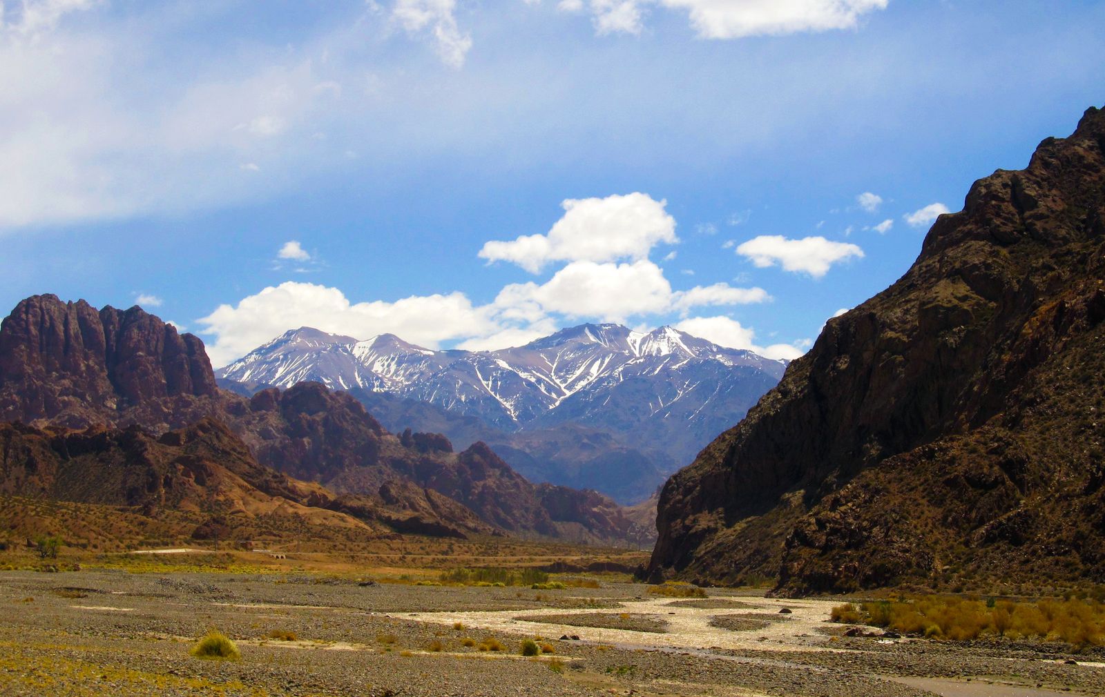 A wide gravel riverbed between red rock walls below a snow-streaked Andean range. Photo by Juan Pablo Lancia on Pexels.