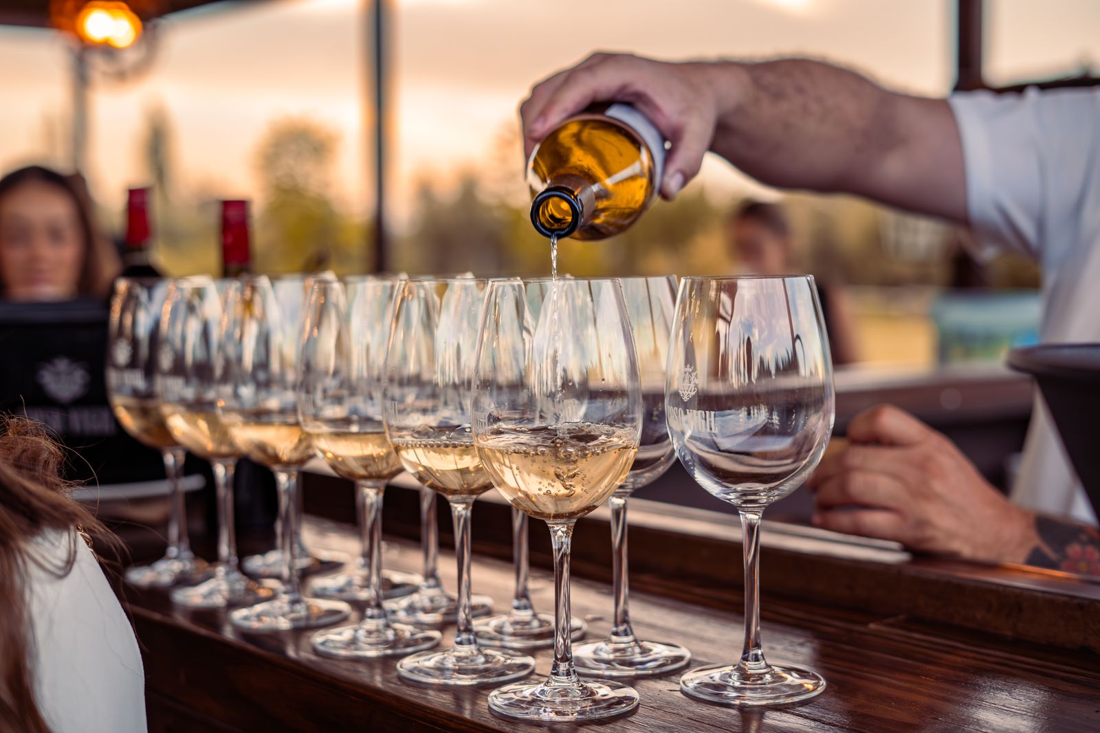 A long row of wine glasses being poured on a bar counter in low evening light. Photo by Nano Erdozain on Pexels.