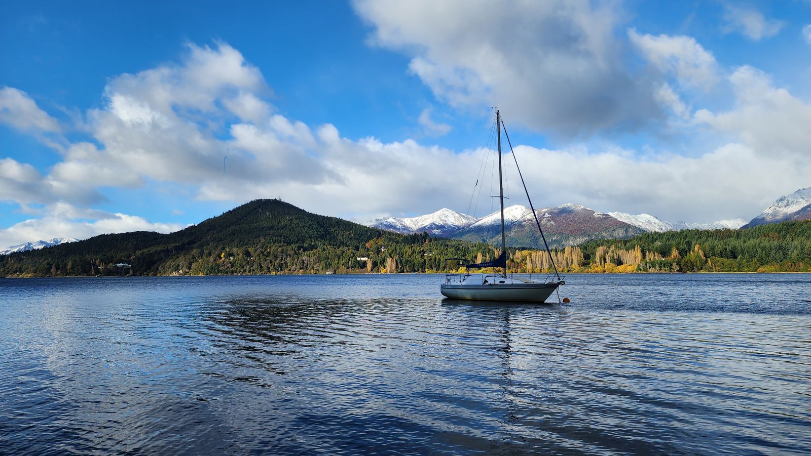A sailing boat anchored on a deep blue lake below forested hills and snow-streaked peaks at Bariloche. Photo by Jazmín Burela on Pexels.