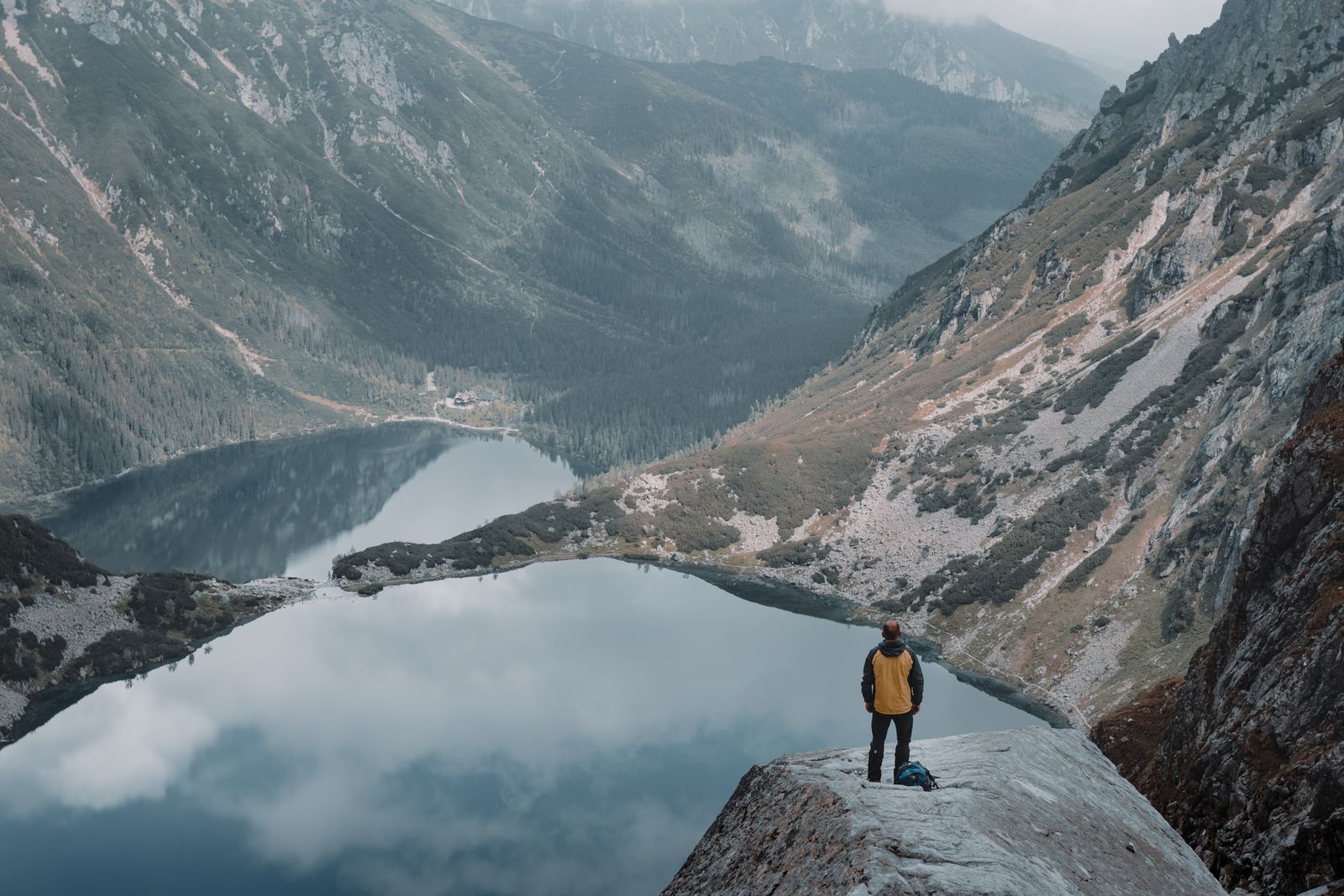 Una persona parada sobre una roca por encima de un lago oscuro y quieto rodeado de laderas grises empinadas. Foto de Gantas Vaičiulėnas en Pexels.