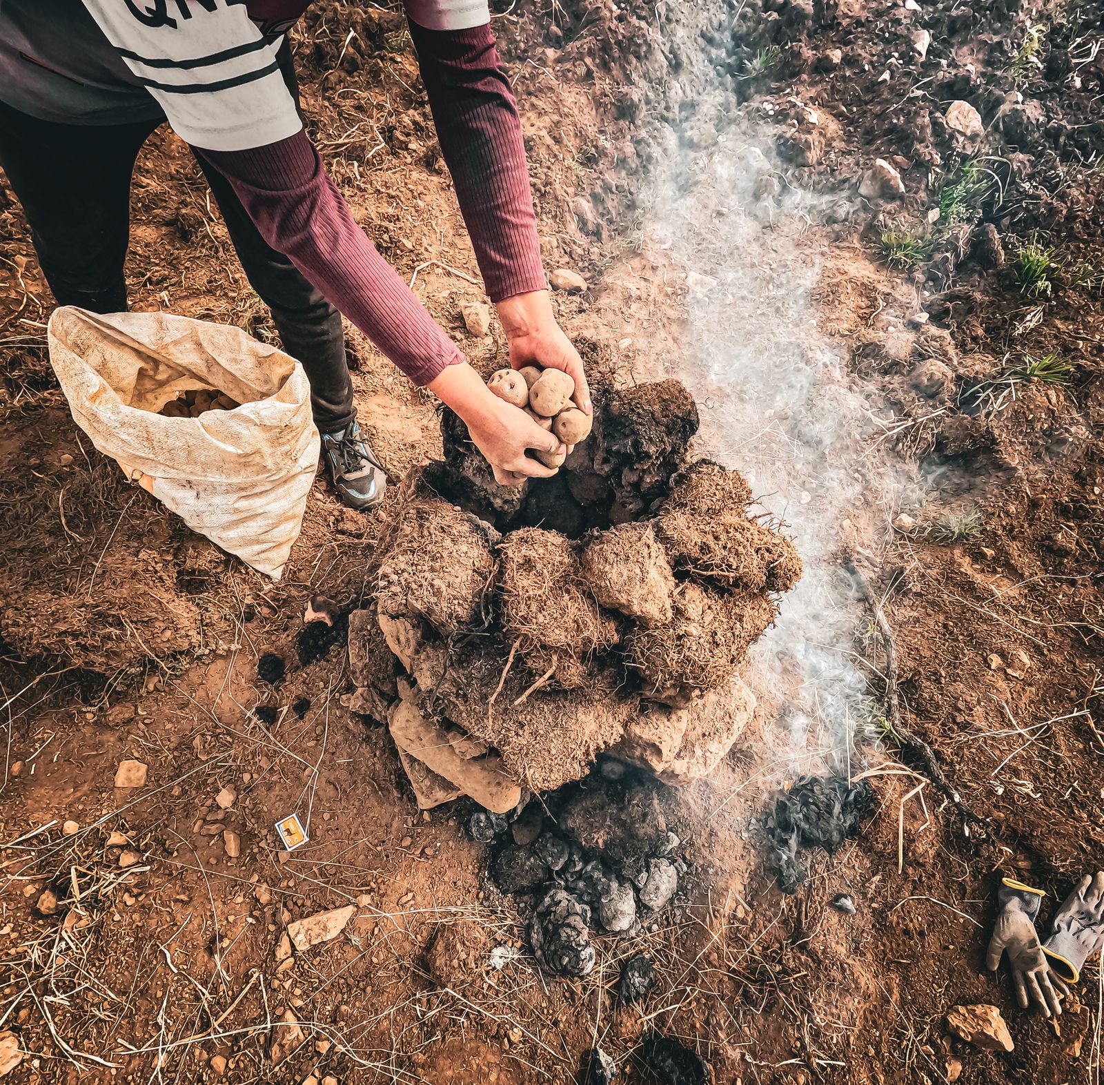 Hands lifting hot stones and earth off a buried oven as the steam comes up. Photo by Luis Dominguez on Pexels.