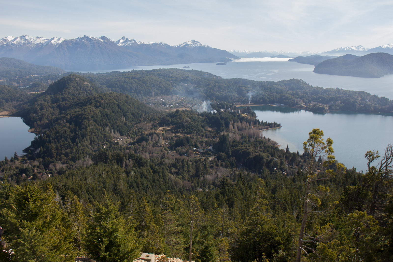 A big lake full of forested peninsulas seen from a wooded viewpoint, with snow-streaked mountains along the far shore. Photo by Pedro Rey on Pexels.