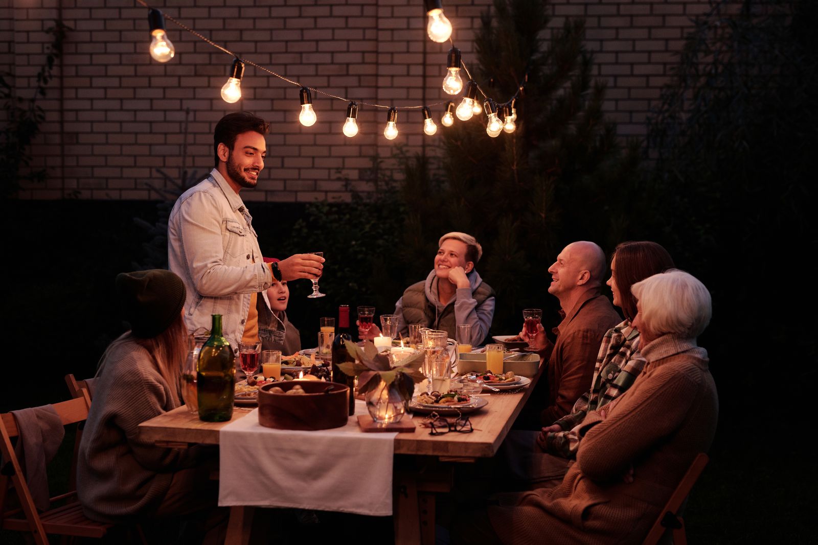 A group of friends at a long outdoor dinner table under string lights. Photo by Askar Abayev on Pexels.