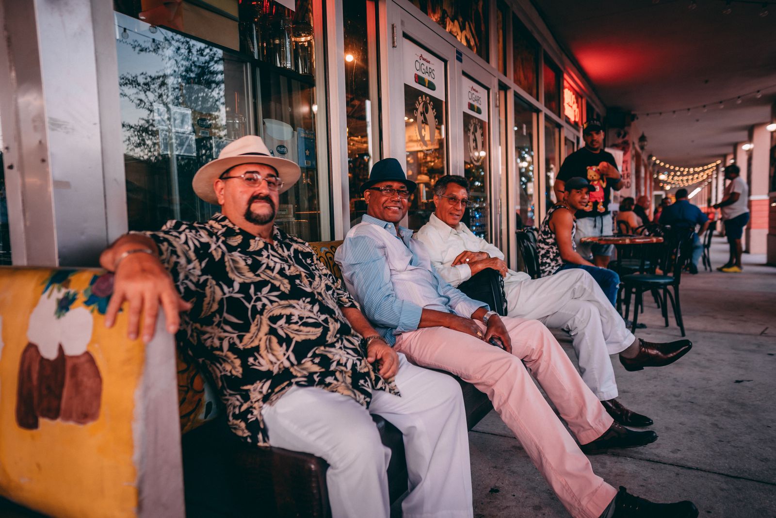 A group of men in hats sitting together outside a lit storefront in the evening. Photo by Sami Abdullah on Pexels.