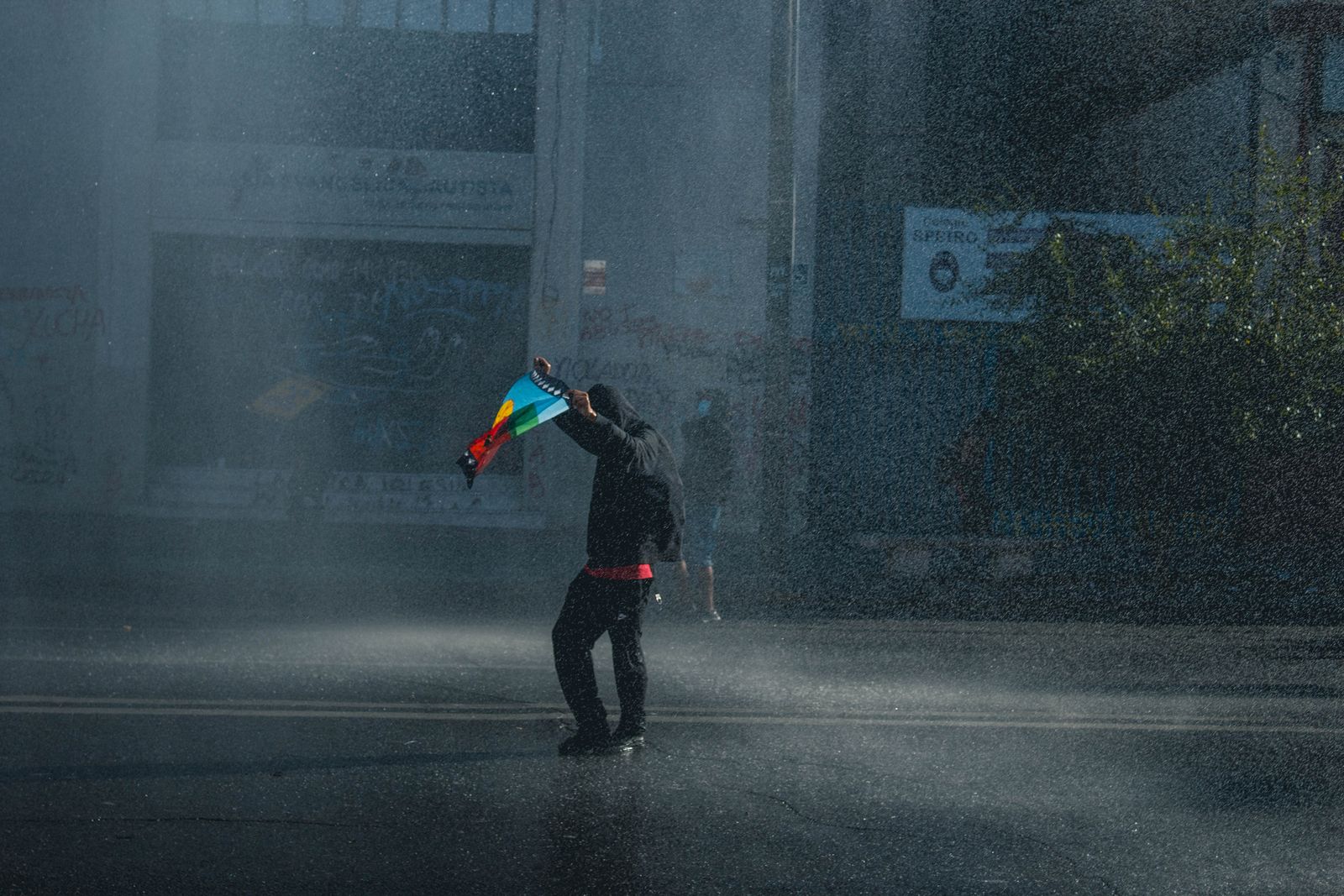 A lone protester holding a flag in the spray of a water cannon on a Santiago street. Photo by Francisco Marín on Pexels.