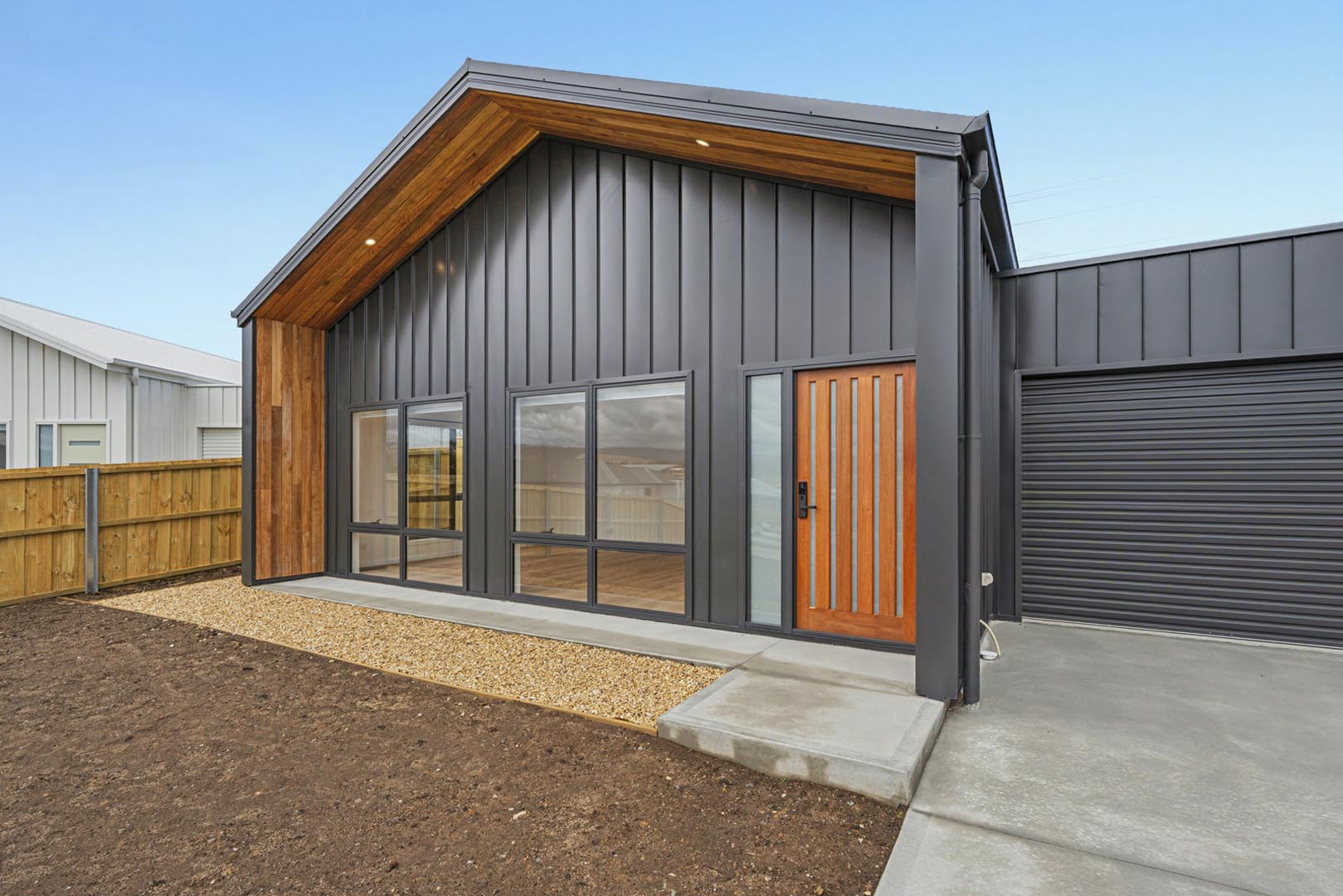 Modern black house with wooden door, large glass windows, gravel path, and concrete driveway.