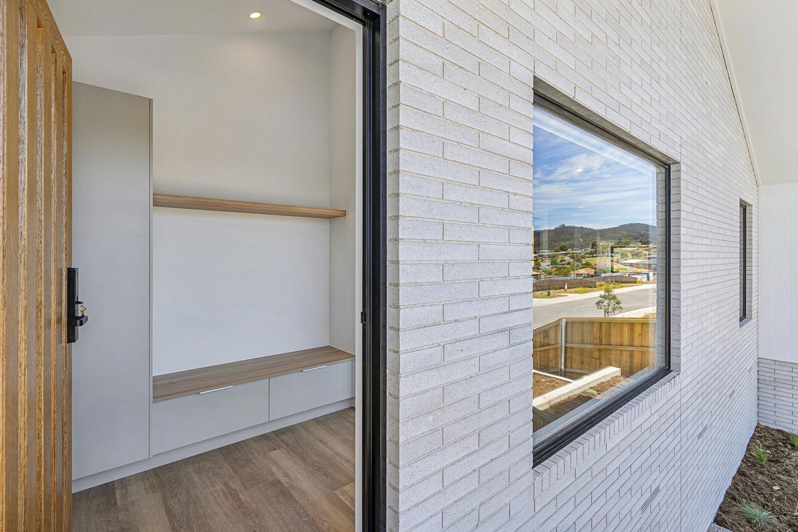 Open wooden door showing hallway with bench, storage cabinets, wooden shelf, and white brick exterior wall.