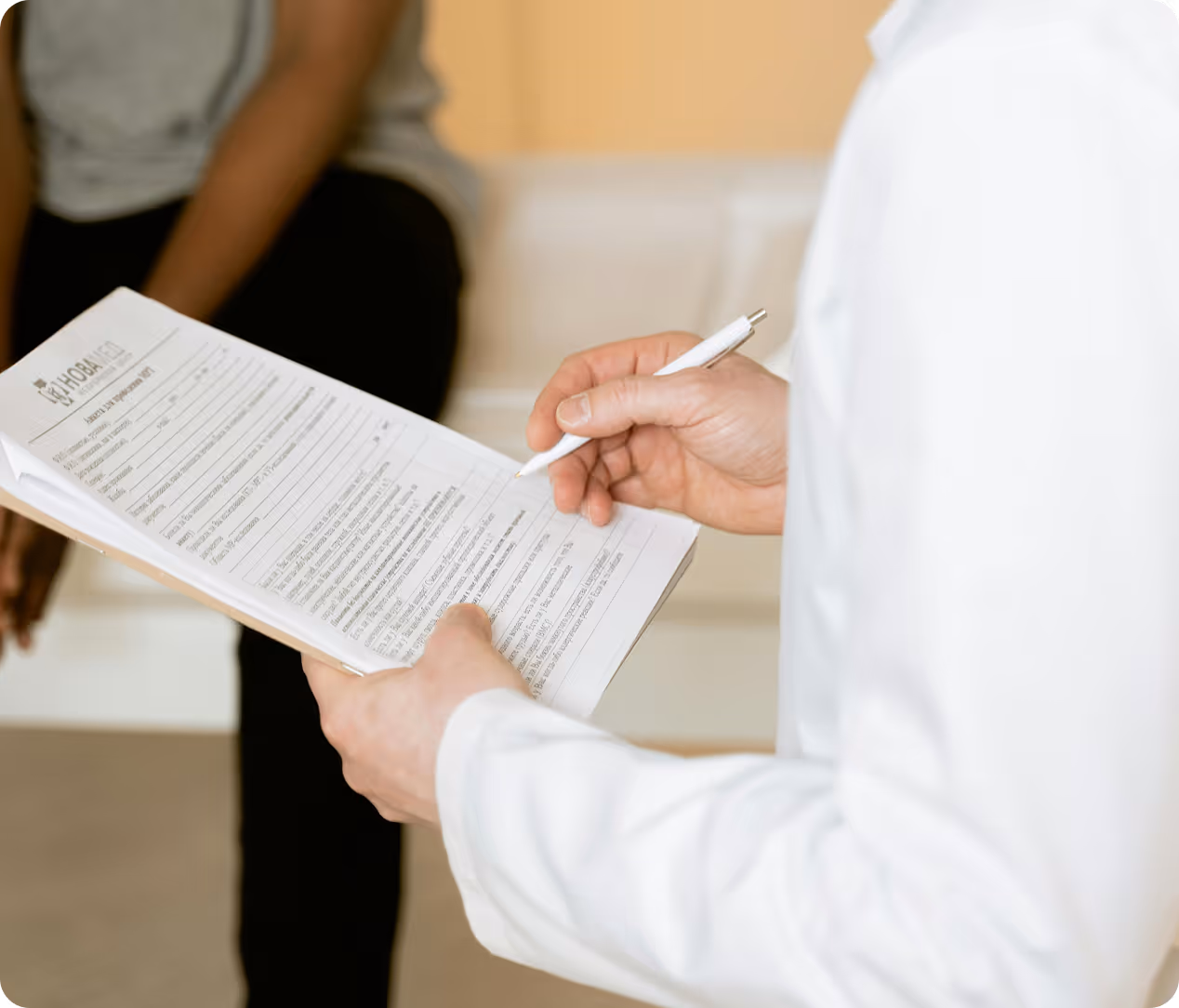 Person in white holds clipboard with medical form, another person sitting in background.