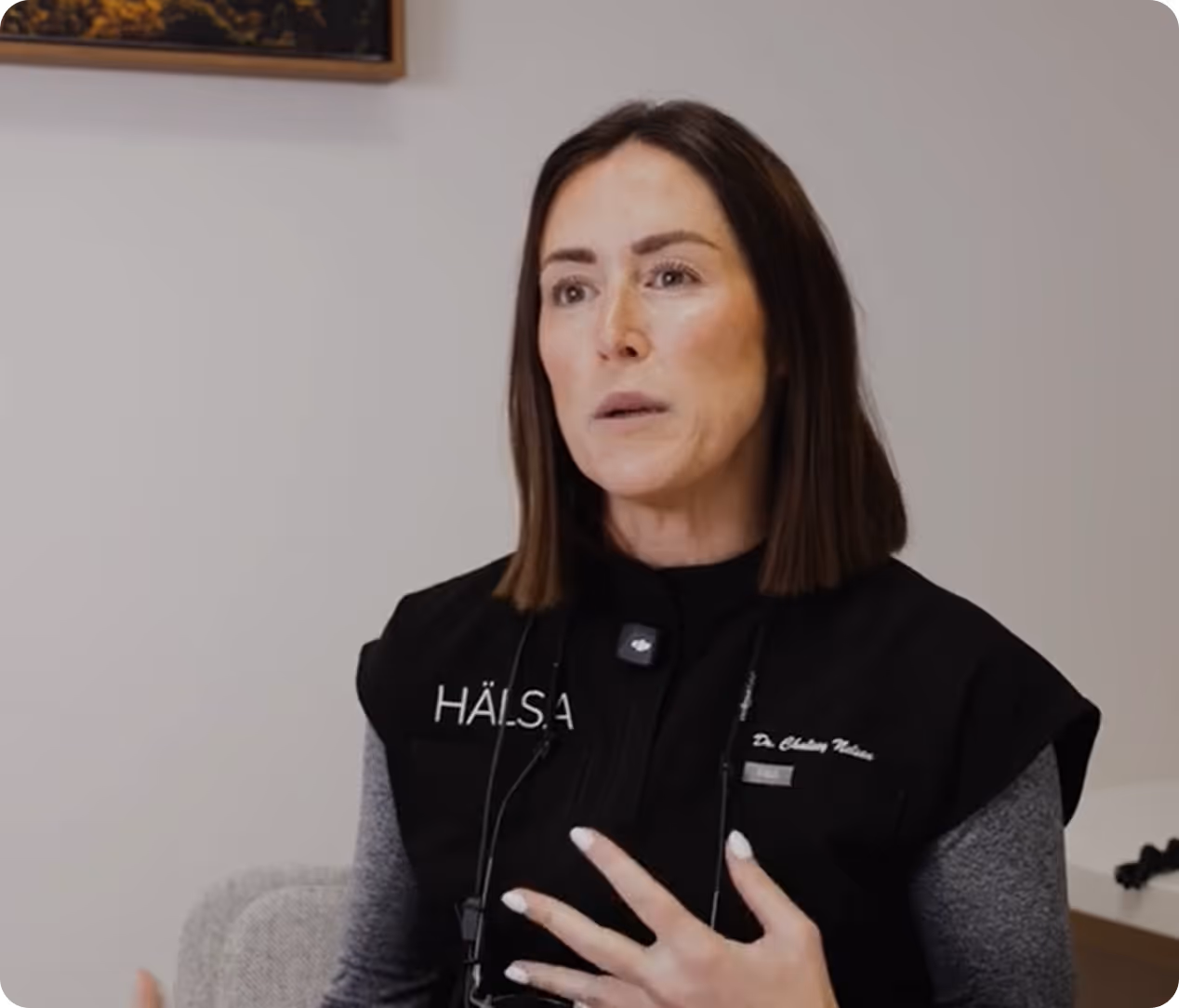 Woman with shoulder-length brown hair speaking, wearing a black vest with 'HÄLSA' and a name tag.