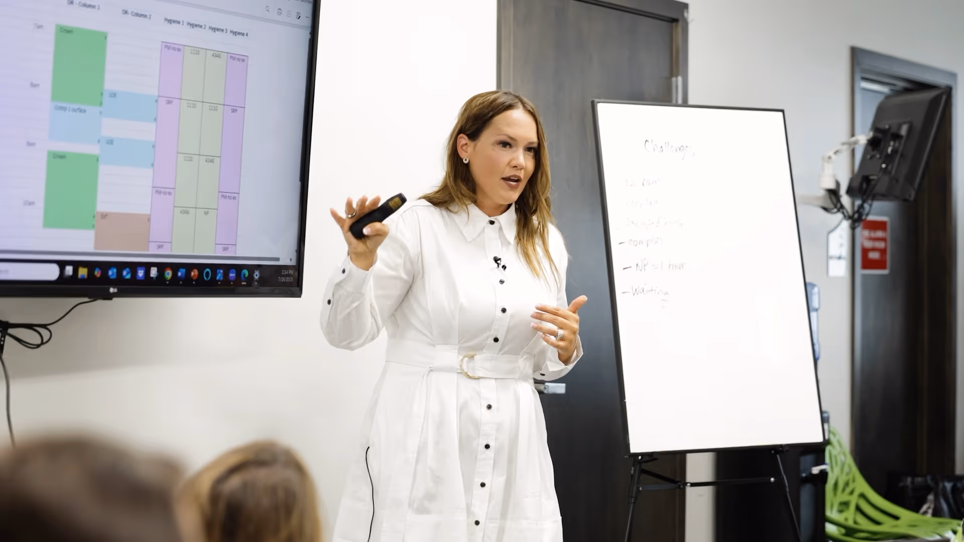 Woman in white dress giving a presentation near a whiteboard and a screen with a schedule chart.