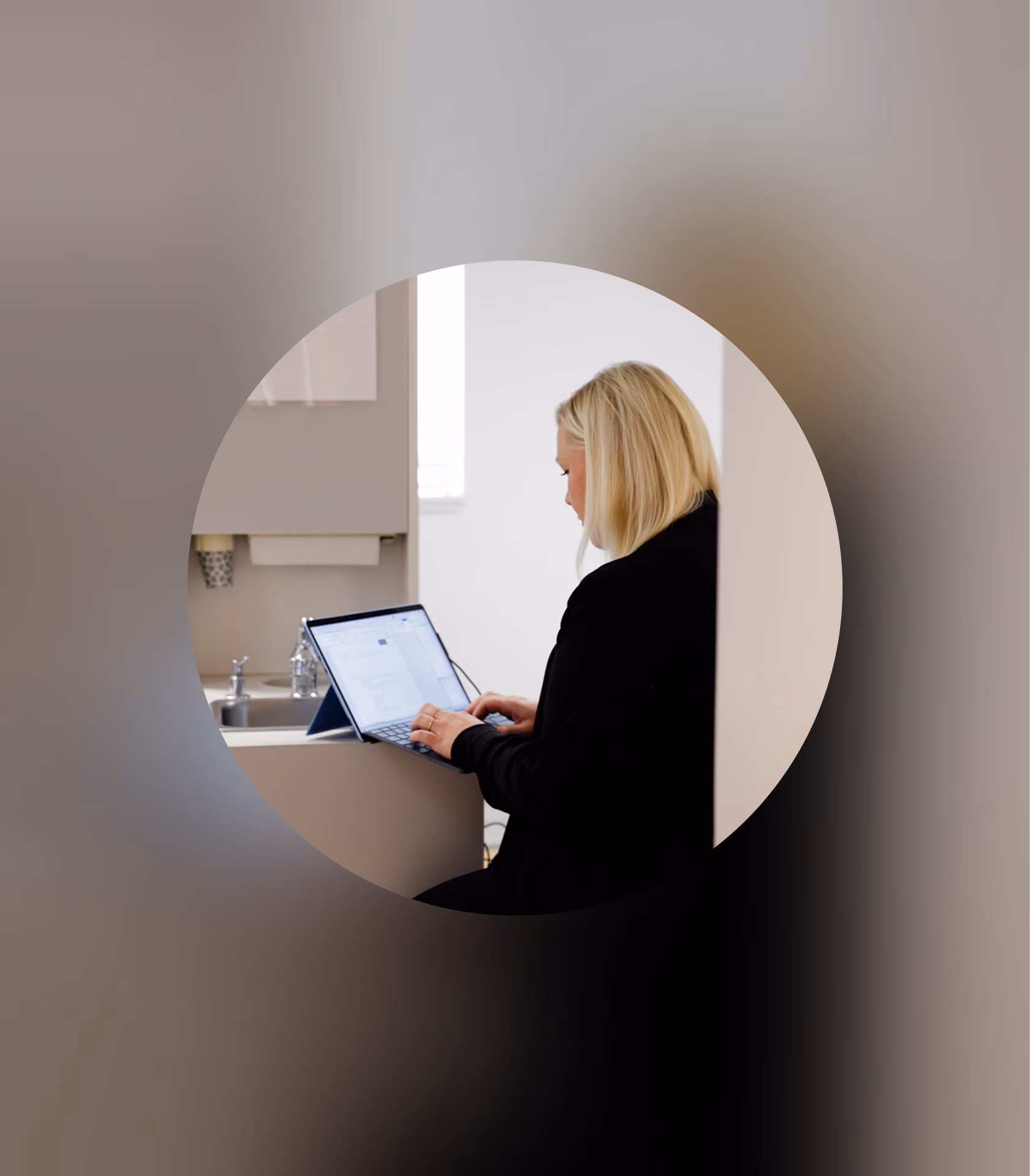 Blonde woman in black working on a laptop in a bright kitchen, seen through a circular frame.