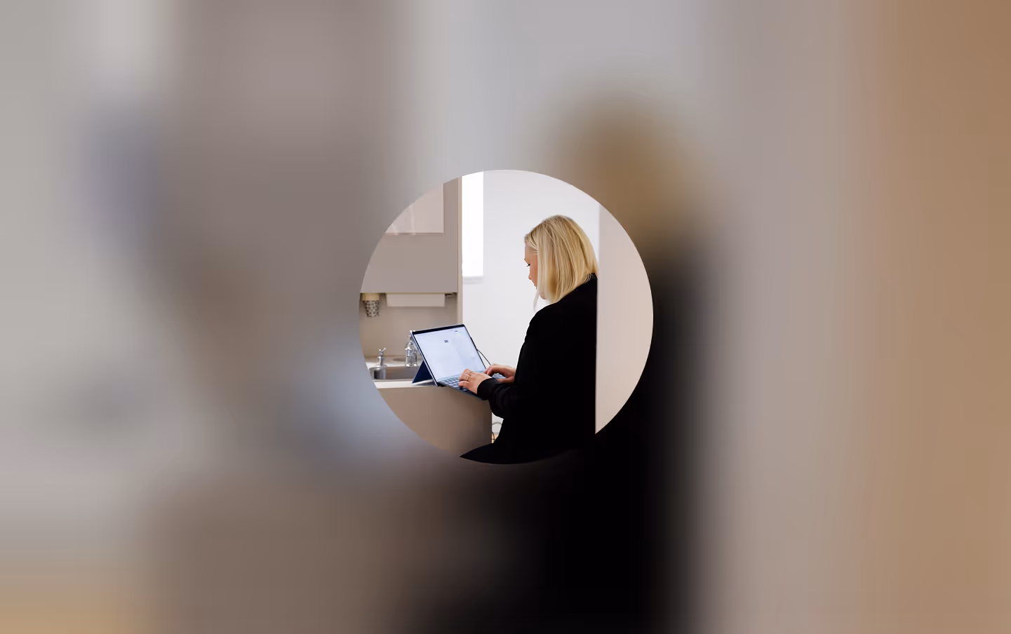 Blonde woman in black working on a laptop in a bright kitchen, seen through a circular frame.