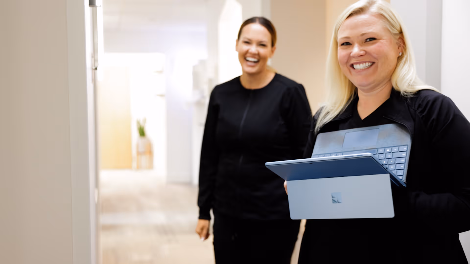 Two women smiling in a hallway, one holding a laptop with a light gray back panel.
