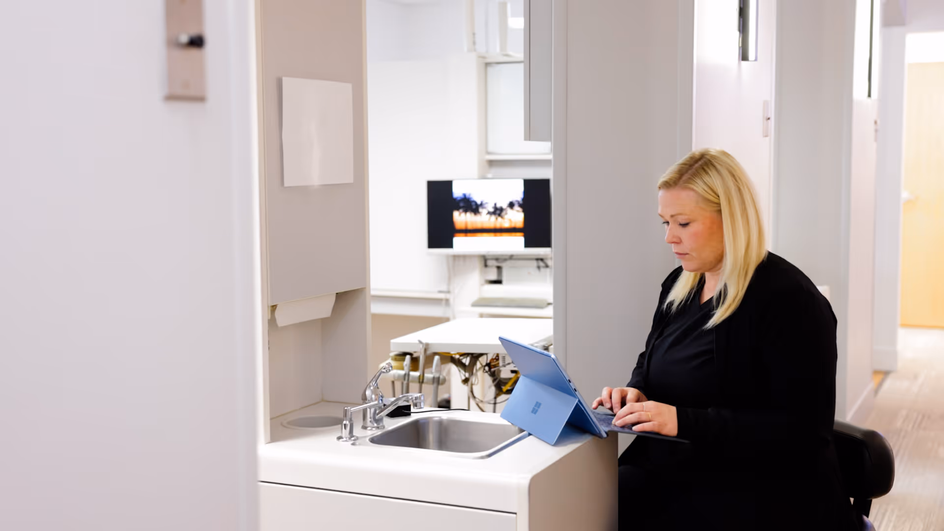 Woman with blonde hair working on a tablet while sitting beside a sink in a bright office space.