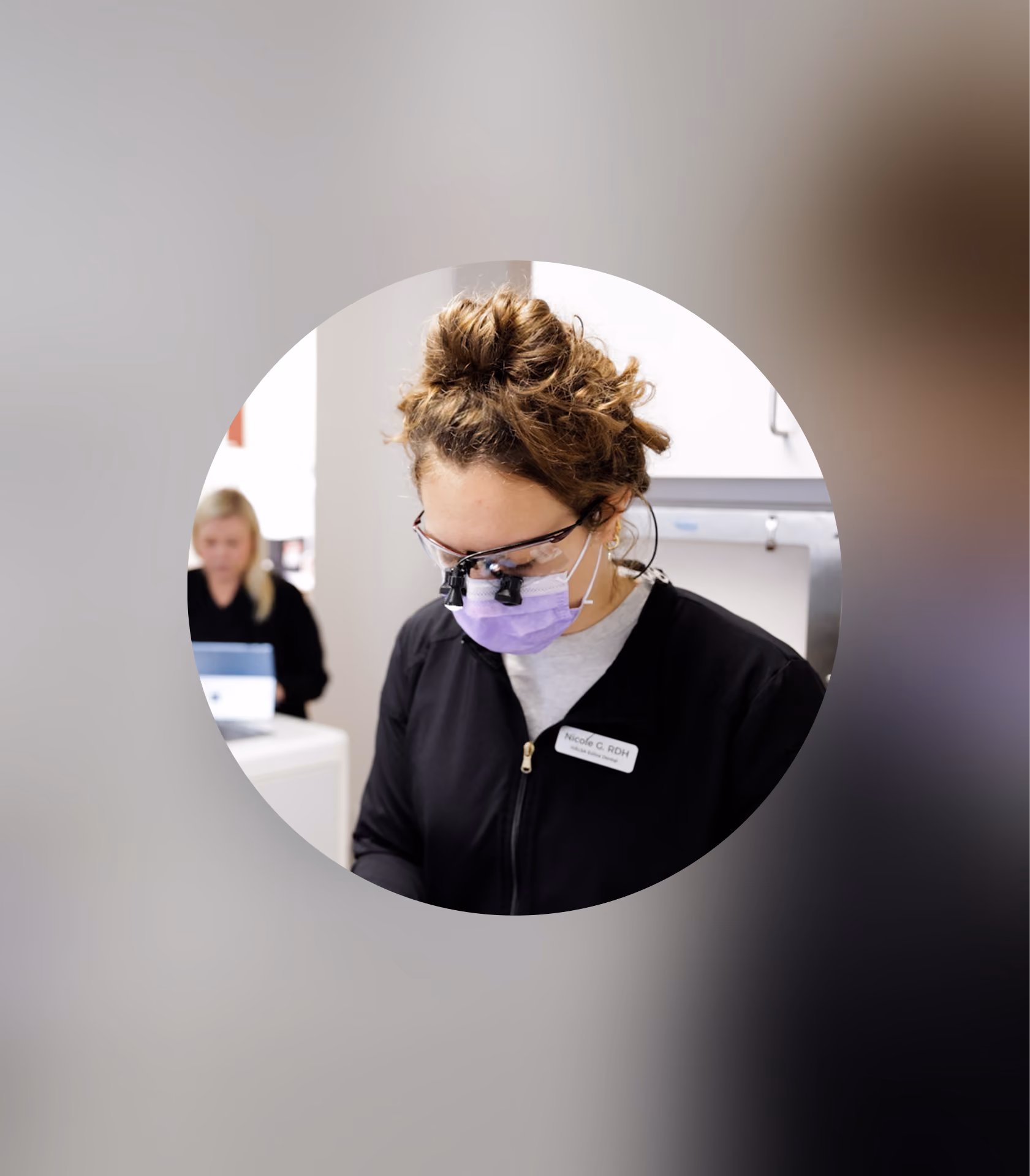 Dental hygienist in black scrubs and purple mask focused on work, with another staff member at desk behind her.