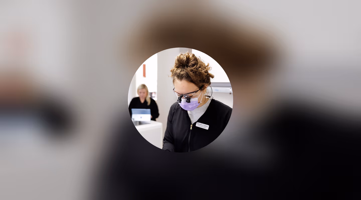 Dental hygienist in black scrubs and purple mask focused on work, with another staff member at desk behind her.